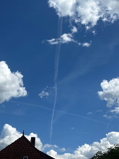 This media file captures a dynamic sky scene with a clear blue expanse dotted with fluffy white cumulus clouds. Several contrails, likely from airplanes, form intersecting lines across the sky, suggesting ongoing air traffic. Two small birds are visible in flight, adding a touch of natural movement to the scene. In the lower right corner, the tiled roof and chimney of a building, characteristic of German architecture, are partially visible, providing a grounded element against the vast sky. The lighting suggests it is daytime, likely late morning or early afternoon, with the sun casting bright illumination on the clouds. The overall mood is one of openness and activity in the sky. No visible text is present in the image. The scene is set in Heinebach, Germany.