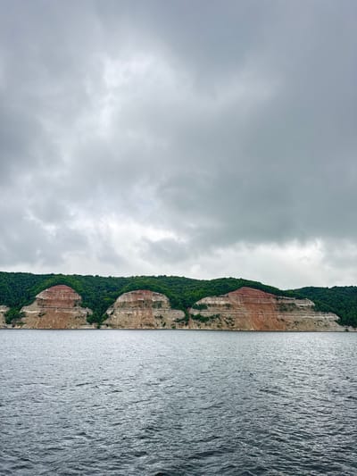 The image captures a panoramic view of a riverbank with steep, layered cliffs in Tenishevo, Russia. The sky is overcast with heavy, grey clouds. The water in the foreground is a dark, choppy grey, indicating a moderate wind. The cliffs rise dramatically from the water's edge, displaying distinct horizontal strata of reddish-brown, tan, and grey rock. The upper portion of the cliffs is covered in dense, dark green foliage, suggesting a forest. The overall scene conveys a sense of natural grandeur and a slightly somber atmosphere due to the cloudy sky.