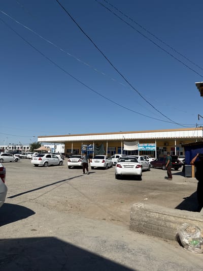 The image depicts a street scene in Pakhtakor, Uzbekistan, under a clear blue sky. The time of day appears to be daytime, with bright sunlight casting distinct shadows.
The primary subjects are several white cars parked along a paved street next to a building. The building has a light-colored facade with a darker trim near the roof. One car has a license plate that reads "05 G 543 AA". Another car's license plate is partially visible, showing "05 U 769 AA". A sign on the building reads "AGRO-NET APTEKA".
There are a few people in the scene, mostly in the background near the building and cars. One person is standing and leaning over the hood of a white car. Another person is walking away from the building. Their attire suggests a casual setting.
The scene appears to be a marketplace or a commercial area, given the presence of shops or businesses lining the street. The overall atmosphere seems calm and ordinary, with no indications of any specific event or heightened emotion. The concrete ground shows some wear and tear, and a plastic bag filled with trash is visible on the side of the road. Overhead, several power lines crisscross the sky.