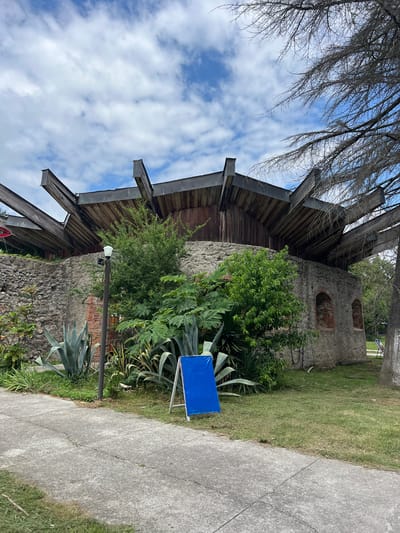 The image displays an outdoor scene with a rustic building constructed of stone and brick. The building features arched openings and a wooden roof structure with exposed beams. Adjacent to the building, there is lush green vegetation, including agave plants and a leafy shrub. A concrete path runs alongside the greenery, and a portion of a grassy area is visible. In the foreground, a bright blue rectangular object, possibly a sign or a panel, is propped up by metal legs. The sky is partly cloudy, with white clouds scattered across a blue expanse. The lighting suggests it is daytime. The overall impression is of a quiet, somewhat weathered outdoor space, possibly a historical site or an area with a unique architectural style. The image appears to be taken from a slightly low angle, looking up towards the building and sky. There is no visible text in the image.