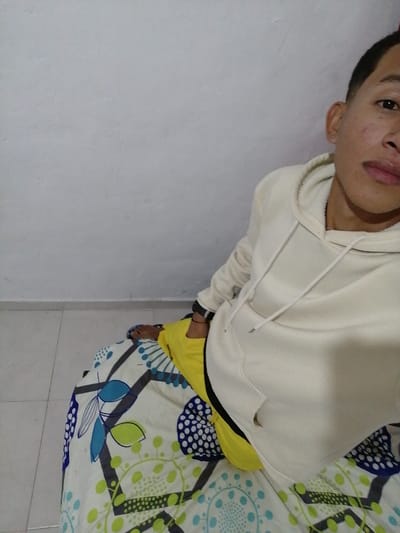 A young person with short dark hair is lying on a bed. They are wearing a light beige hoodie and yellow pants. The bed has a patterned sheet with blue, white, green, and black designs. A white wall is visible behind them. The person is facing slightly towards the camera and the angle suggests a selfie. The room is lit by a soft light.