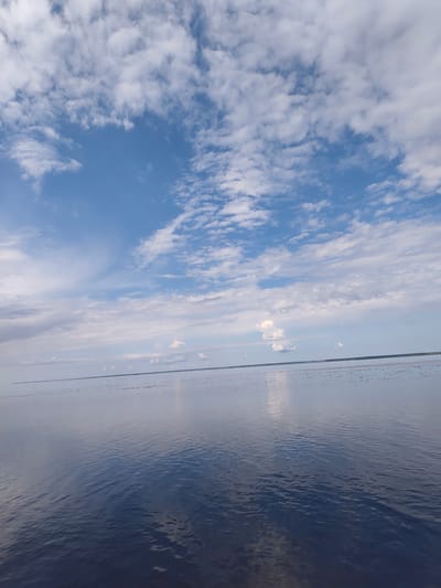 The image depicts a wide expanse of calm water under a bright blue sky filled with scattered white clouds. The horizon line is clearly visible, separating the water from the sky. The water's surface is smooth and reflective, mirroring the sky and clouds. The far distance shows a faint, dark line, likely the coastline or a low-lying landmass. The overall scene is serene and expansive, suggesting an open body of water such as a lake or sea, possibly in a rural or natural setting outside of a densely populated area. The location is identified as Kosachivka, Ukraine.