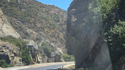 A two-lane highway cuts through a mountain pass. The road is flanked by steep, rocky slopes covered with sparse vegetation. A massive, gray rock formation looms on the right, with trees growing near its base. Sunlight illuminates the road and parts of the rock face. The sky above is clear and blue.