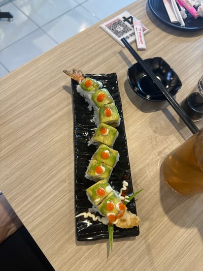 This image captures a top-down view of a dining setting, likely in a casual restaurant in Kecamatan Cisaat, Indonesia. The main subject is a long, black rectangular plate holding an attractively presented sushi roll. The sushi appears to be a "dragon roll" or similar, featuring thin slices of avocado on top, adorned with small dots of orange roe or sauce, and a decorative drizzle of white creamy sauce. The left end of the roll clearly shows the tail of a fried shrimp tempura.
On the light wood-grained table, a pair of black chopsticks rests diagonally on a small black, diamond-shaped dish, likely for soy sauce. To the left, a small black plate contains discarded napkins or wrappers. A white card with a large QR code and the visible text "ONE-SAN" (potentially preceded by Japanese characters) is also present, likely serving as a menu or payment prompt. A dark-colored bottle, possibly soy sauce or chili oil, is partially visible in the upper left. A tall glass filled with an amber-colored iced beverage, such as iced tea, is seen in the upper right. The scene depicts a meal laid out, ready for dining. Part of a person's leg or clothing is visible in the bottom right, suggesting someone is seated at the table.
