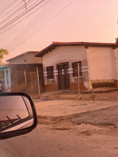A photograph captures a static street scene in La Cruz del Pastel, Venezuela, under a warm, pale orange or pink sky, indicative of early morning or late afternoon. The image is taken from inside a vehicle, with the black frame of a side mirror visible in the lower right foreground, reflecting utility wires and possibly parts of the surrounding structures.

The central focus is a single-story house with a prominent red tiled roof and light-colored walls, possibly painted white or light beige. Several windows are visible on the front facade, each protected by dark-colored security bars arranged in a grid pattern. A section of chain-link fence or metal mesh runs along the front of the property, partially obscuring the view of the house and its immediate surroundings. Below the main house, another lower structure, possibly an extension or a separate building, has darker, unpainted walls and a distinct light blue lower section.

The ground in front of the house appears to be a mix of dirt and uneven concrete, suggesting an unpaved or partially paved road or pathway. Power lines or utility wires crisscross the upper left portion of the sky, adding to the visual texture of the scene. A small amount of green foliage is visible near the bottom left, hinting at some vegetation in the area. No people are visible, and there is no discernible activity taking place. The overall impression is that of a quiet residential area. No text is visible in the image.