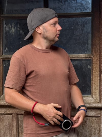 A man in a brown t-shirt and grey baseball cap is positioned in front of what appears to be an old wooden window frame. He holds a black smartphone with a large circular camera lens. The man has a dark watch and beaded bracelet on his left wrist and a red bracelet on his right. The man's face is tilted slightly up. The window behind him contains dark glass, possibly reflecting or obscured. The image is rotated; it would appear the man is lying down. The location of Sà Phìn Commune, Vietnam is not explicitly identifiable in the image.