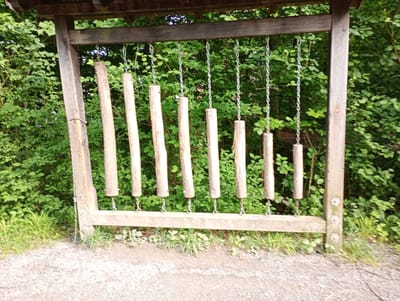 An outdoor wooden instrument, resembling a xylophone with wooden bars, is suspended by chains from a wooden frame. The instrument is set against a backdrop of lush green trees and foliage. The ground in front of the instrument is covered in gravel. The scene appears to be in a park or natural setting. This outdoor musical instrument is located in the community center of Brannenburg, Germany.