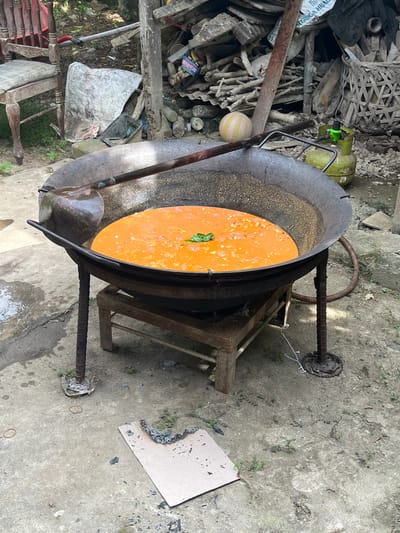 In Keudematangkuli, Indonesia, a large, well-used wok sits over a makeshift fire pit, its contents a bubbling, orange-hued stew or curry. The wok is supported by a sturdy, rustic metal stand fashioned from rebar. A large ladle rests within the wok, hinting at recent or ongoing cooking. Surrounding the cooking area, the environment appears to be an outdoor space with a concrete floor, showing signs of recent use with scattered debris, including burnt cardboard and a rubber band. In the background, a pile of salvaged materials, including wood, corrugated metal, and what appears to be a gas cylinder with a green regulator, suggests an area used for practical purposes, possibly a backyard or workshop. A traditional wooden chair with red detailing is visible to the left. The overall impression is one of resourcefulness and everyday life in a rural or semi-rural Indonesian setting.