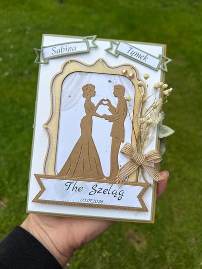 A handmade wedding card is held by a person's hand. The card features a silhouette of a bride and groom forming a heart shape with their hands. Above the couple, two banners read "Sabina" and "Tymek" in green text. Below the couple, another banner reads "The Szelag" with the date "03.07.2026" underneath. The card is decorated with dried flowers and a burlap bow. The background is a grassy area, suggesting an outdoor setting. The location of this image is not Belfast City, United Kingdom.