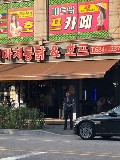 The image shows a street scene in Pyeongtaek-si, South Korea, featuring a storefront with large advertisements. The storefront appears to be a restaurant or cafe, with a brown awning extending over the entrance. Above the awning, two large banners display Vietnamese text and images of women in traditional Vietnamese attire. The left banner features the text "Viet Nam HOF & Quán cà phê" and a woman holding a conical hat and flowers. The right banner has "2F 소주.맥주.양주" and an image of a woman wearing a conical hat. Below these banners, a red sign displays Korean text, likely the name of the establishment, "마치통닭 & 호프", and a phone number, "T.684-1237".
In front of the establishment, a man wearing sunglasses, a patterned vest over a dark shirt, and dark pants stands smoking a cigarette. To the right, a black car is parked, partially obscuring the view of the building's interior. The interior appears dimly lit with people visible inside. The street in the foreground is paved and shows clear lane markings. The overall setting is a commercial area during daylight hours.