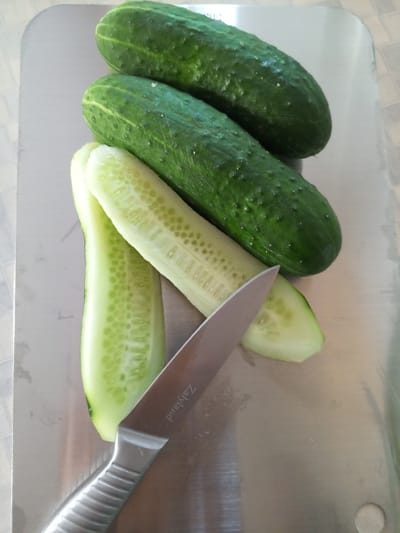 This is a close-up, overhead shot of three whole cucumbers and one partially cut cucumber on a stainless steel surface. The cucumbers are dark green and bumpy. One cucumber has been cut lengthwise, revealing its pale green interior and seeds. A silver knife with a black handle is positioned diagonally across the bottom of the image, with its blade touching one of the cut cucumber halves. The knife has the word "Zalyand" etched into its blade. The background is a light-colored, tiled surface. There is no indication of the city of Vilyuysk, Russia, in the image.