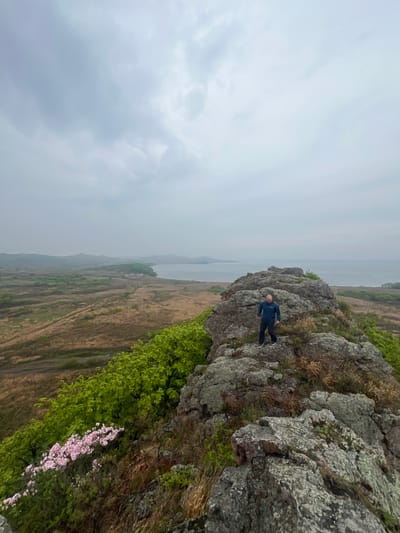 A wide, slightly tilted, outdoor shot shows a man in dark clothing standing on a rocky outcropping. The environment is overcast, with the sky taking up the majority of the left portion of the image. In the center of the image, a body of water extends into the distance. The ground below the rocks is a mix of brown fields, green vegetation, and patches of pink blossoms. The man, bald, is wearing a blue jacket and dark pants. He stands on the edge of the rock formation, facing toward the center of the frame. The shot may have been taken near Gvozdevo, Russia.