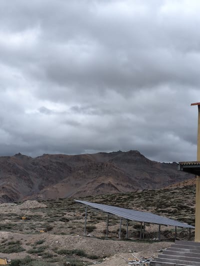 A solar panel array is positioned in the foreground of a rugged, mountainous landscape under a cloudy sky. The mountains are barren and appear to be made of rock and soil, with layered geological formations visible. Sparse vegetation, low-lying shrubs and grasses, dots the ground around the solar panels. To the right, a portion of a building with a yellow facade and a concrete staircase leading up to it is visible. The sky is filled with heavy, gray clouds, suggesting overcast weather. The overall scene depicts a somewhat remote, possibly arid, environment where renewable energy infrastructure has been installed. There are no people or animals visible in the image. The location, Dhar Chharp Samba, India, suggests a high-altitude, potentially mountainous region.