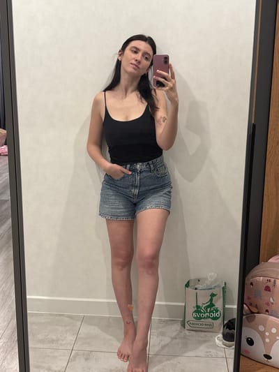 A young woman poses for a mirror selfie. She wears a black tank top and denim shorts, her left hand tucked in her pocket, and holds a rose-colored phone with her right hand. The mirror reflects a glimpse of a backpack with an animal print in the top right corner. On the floor, a shopping bag stands next to discarded shoes. A small butterfly sticker adorns her lower leg.