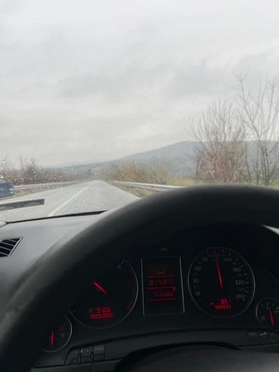 The image presents a first-person perspective from the driver's seat of a vehicle in the vicinity of Aleko Konstantinovo, Bulgaria, on an overcast day. The time displayed on the vehicle's instrument cluster is 9:08 on March 18, 2026.
The windshield is covered in numerous raindrops, indicating precipitation. Ahead, a two-lane asphalt road, marked by a dashed white center line and a solid white right edge line, stretches into the distance, appearing wet. A metallic guardrail runs along the right shoulder of the road. Beyond the guardrail, bare trees and patches of green vegetation are visible. A distant range of hills or mountains is visible under a uniformly grey, cloudy sky. A portion of a light-colored vehicle is discernible on the far left side of the road.
The vehicle's dark dashboard and instrument cluster occupy the lower half of the frame. The speedometer indicates a speed of 89.1 km/h, with an odometer reading of 368,685. The tachometer shows the engine operating at approximately 1,000 RPM. A central digital display shows an ambient temperature of +4.5°C and an average fuel consumption of 6.2 L/100km. An illuminated warning symbol, appearing as a radiator/cooler with a snowflake, is present on this display. The fuel gauge indicates approximately a quarter tank remaining, and the engine temperature gauge is positioned at a low operational reading. No individuals are visible in the scene.