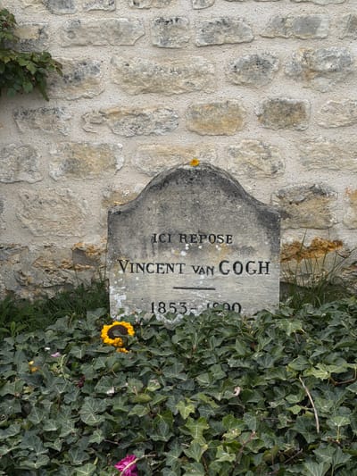 The image depicts the tombstone of Vincent van Gogh, located in the cemetery of Auvers-sur-Oise, France. The gray stone marker is partially overgrown with ivy and set against a light-colored stone wall. The inscription reads "Ici Repose Vincent van Gogh 1853-1890" vertically. Two knitted sunflowers and a wilting pink flower lie among the ivy at the base of the tombstone.