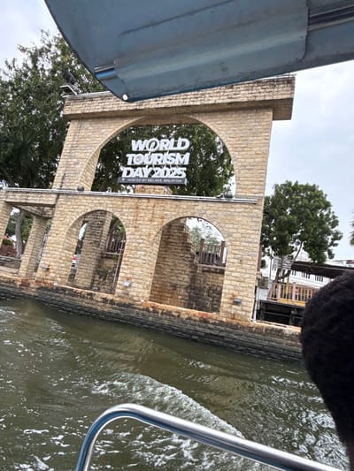 A partially obscured view from a boat captures an aged, stone structure with multiple arches. A sign displaying "World Tourism Day 2025" adorns one of the arches. The structure borders a waterway, with trees and building elements visible in the background. The boat's components are also present in the shot, with the water showing ripples from its movement.