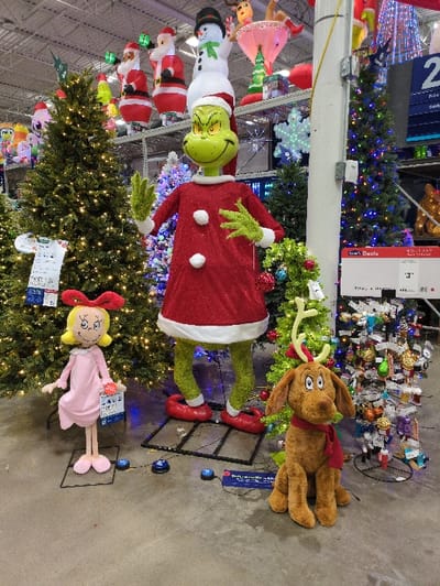 A retail display features holiday merchandise. A large Grinch figure, dressed in a red coat, stands amidst Christmas trees. Cindy Lou Who and Max, the Grinch's dog, are positioned near the trees. Ornaments and lights adorn the trees. Santa Claus figures hang from the ceiling, adding to the festive atmosphere. The scene is set in a store environment.