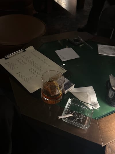 The image depicts an interior scene at a dimly lit, high-end bar or lounge in Guadalajara, Mexico. The primary subject is a dark-edged, green-felt table surface.

On the table, a white paper menu secured on a silver clipboard is positioned on the left, displaying items in Spanish with visible sections including "LICORES," "MEZCAL," "GINEBRA," and "BOTELLAS." Adjacent to the menu, a clear rocks glass contains an amber liquid, large square ice cubes, and a lemon or orange peel garnish. To the right of the glass, a dark-colored pack of cigarettes is partially visible. Further right, a clear glass square ashtray holds cigarette ash and one unlit cigarette resting on its edge.

Several folded white paper napkins are scattered across the table; one napkin prominently displays the handwritten text "ALBANTO DUMBO," and another appears to have a partial handwritten note. A light-colored smartphone, positioned face down, and a slender pen rest in the upper-center area of the table.

In the background, out of focus due to shallow depth of field, are dark furniture elements, including a curved chair back on the left and a dark-clad leg and foot visible on the right, indicating the presence of at least one individual seated or standing nearby. The overall ambiance is subdued and refined, characteristic of an upscale establishment.