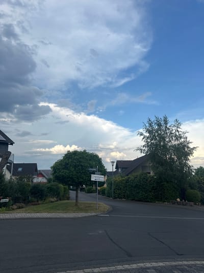 The image depicts a street intersection in Sohren, Germany, under a sky with mixed clouds and blue patches. To the left, a grey slate-roofed house with a balcony is visible, adjacent to a smaller house with a red roof and dark windows. A dark green, round-topped tree stands in the center of the intersection, near a street sign that reads "Breitenacker" and points to "Praxis für Physiotherapie" (Physiotherapy Practice). Further to the right, a tall, bushy tree with sparse foliage dominates the scene, partially obscuring a building with a dark tiled roof and beige siding. Dense green hedges and other trees line the properties. The road is paved with asphalt and bordered by cobblestones. A street lamp is positioned to the left of the large tree. The environment appears to be a residential area.