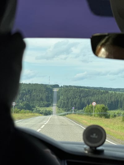 The image depicts a view from inside a vehicle driving on a paved road towards a distant horizon under a cloudy sky. The road is lined with green trees and grass, with rolling hills in the background. There are two traffic signs visible on the right side of the road: a circular sign with a red border and two red cars depicted, indicating "no overtaking," and a blue circular sign with a red border and a diagonal red line, likely a prohibition sign. The car appears to be traveling in a rural area. The scene is likely in or near the village of Golyany, Russia, given the style of the road and surrounding landscape, and the presence of typical Russian road signage. A blurry silhouette of a person's head and shoulder is visible in the foreground on the left, suggesting the photographer is a passenger or driver. A car accessory, possibly a phone mount or GPS device, is visible on the dashboard in the lower right corner.