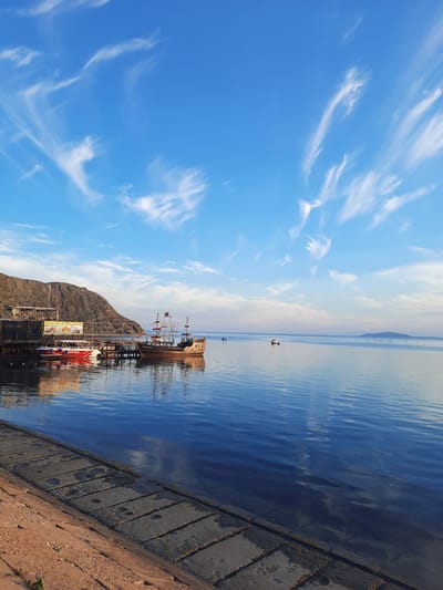 The image depicts a serene waterside scene, likely in the city of Ordzhonikidzevsky Settlement Council, Ukraine, given the provided context. The foreground features a concrete embankment leading to calm, deep blue water that reflects the sky and surrounding objects. On the left, a rocky cliff rises, with a wooden pier extending into the water. Two boats are docked at the pier: a smaller red and white speedboat and a larger, older-looking wooden ship, resembling a pirate ship, with masts and rigging. The water is still, with gentle ripples. In the background, the horizon is marked by a soft, pale sky with wispy white clouds scattered across a clear blue expanse. A distant island or landmass is visible on the far right horizon. The overall atmosphere is peaceful and tranquil, suggesting a calm day at the harbor or waterfront. There are no people visible in the image.
