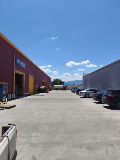 The image shows an outdoor industrial area on a clear, sunny day with scattered clouds. In the foreground, a portion of a white pickup truck is visible on the left. The middle ground features a wide, grey concrete lot. On the left side of the lot, a large, burgundy warehouse-like building with a yellow trim and two large yellow doors is prominent. A sign on this building reads "АРТОАЗА" and "artoaza.com". Several air conditioning units are mounted on the wall of this building, and stacks of wooden pallets are visible in front of it. Further back on the left, a white tanker truck is parked.
On the right side of the concrete lot, a row of parked cars is visible, including a black SUV, a dark sedan, a silver sedan, and a dark hatchback. Beyond the parked cars, another large building with a light grey facade runs parallel to the parked vehicles. In the background, trees and distant mountains are visible. The overall scene depicts a commercial or industrial storage and logistics area.