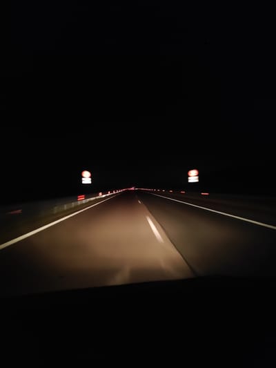 The media file depicts a nighttime view from inside a vehicle driving on a highway. The scene is dark, illuminated primarily by the vehicle's headlights, which cast a warm glow on the asphalt road ahead. White lane markings guide the way. On the right side of the road, a series of red reflectors are visible, suggesting a barrier or edge. Further down the road, a sign with a red circle and a diagonal line through it, likely indicating a speed limit restriction, is illuminated. Beyond that, more red lights are visible in the distance, hinting at other vehicles or illuminated structures. The overall impression is of a solitary journey on a dark highway. The time of day is clearly night. There are no visible people, and the weather appears clear. The location is identified as Kozare, Bulgaria.