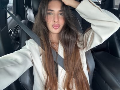 The image shows a young woman with long, light brown hair wearing a white blazer. She is in a car, wearing a seatbelt. The interior of the car has black leather seats. The image was taken indoors. It is not possible to verify the location of North Miami Beach, United States of America.