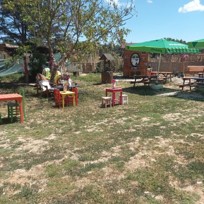 In Çatalca, Turkey, this image captures an outdoor seating area with children's furniture, picnic tables, and large green umbrellas. A wooden structure resembling a small house or playhouse is visible, featuring a round window and decorative elements. Various trees and foliage surround the area, suggesting a park-like or garden setting. The ground is a mixture of dry earth and sparse green grass. Several wooden picnic tables with benches are set up, some under umbrellas, for outdoor dining or gathering. Colorful children's tables and chairs, made of wood, are also present, hinting at a family-friendly environment. In the background, a fence and more trees contribute to the natural, rustic ambiance. A small green toy tractor is parked near one of the picnic tables. The sky is blue with scattered clouds, indicating a clear day.