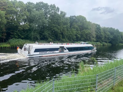 A white passenger boat named "BELVEDERE" is traveling on a dark, calm river. The boat has a blue stripe along its side and the words "SCHIFFFAHRT in POTSDAM .de" are written on it. Several passengers are visible on the upper deck. The riverbanks are lined with tall, green trees and reeds. A metal fence is in the foreground, with tall grass growing behind it. The sky is overcast. The boat is heading away from the viewer towards the right side of the image. The image appears to have been taken in Teltow, Germany, as indicated by the text on the boat.