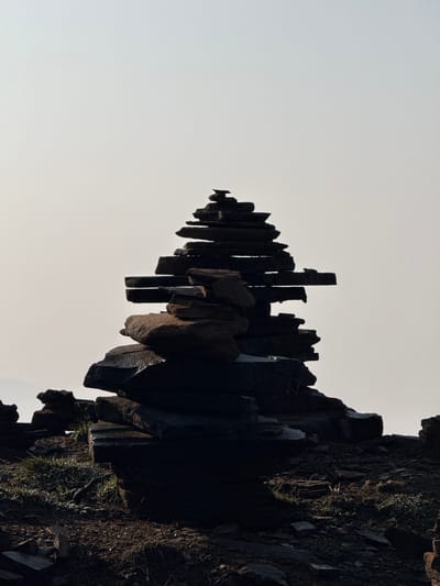 The image, which appears to be rotated 90 degrees clockwise from its natural orientation, captures a dramatic natural landscape featuring a prominent dark rock formation against a light, clear sky. When viewed in its likely natural orientation (rotated 90 degrees counter-clockwise), the formation consists of numerous tall, dark, and jagged columnar rock pillars or spires, resembling a natural fence or a row of ancient ruins.
In the provided orientation, this striking formation extends horizontally from the bottom-right corner of the frame towards the top-left, appearing as a long, uneven crest. The individual rock elements are sharply silhouetted in deep shadow against a vast, bright, clear, light grey sky that dominates the upper-left two-thirds of the image. The pillars vary in height and thickness, creating a highly textured and uneven profile.
Towards the bottom-right of the image, the rock formation widens into a more massive cliff face, where rough textures, fragmented surfaces, and patches of green vegetation and moss are discernible, indicating natural growth on the rock. The strong contrast between the deeply shadowed rocks and the luminous sky indicates powerful backlighting, characteristic of a sunrise or sunset, or a bright day with the structures casting long shadows.
There are no human subjects, man-made objects, or specific activities depicted. The scene conveys a sense of rugged natural beauty, geological grandeur, and an imposing scale. No text is visible. The setting is an outdoor, mountainous or rocky environment in Lwang Ghalel, Nepal.