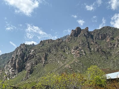 The image captures a rugged mountain landscape in Agarak, Armenia, under a blue sky with scattered white clouds. The mountain is characterized by a mix of rocky terrain and sparse vegetation, with shades of brown and green. Boulders and cliffs are visible throughout the slope. In the foreground, there are trees with light green leaves, partially obscuring small structures and a building with a light blue roof. The composition emphasizes the scale and natural features of the mountainous terrain. A cross-shaped structure is perched at the top of one of the mountain peaks.