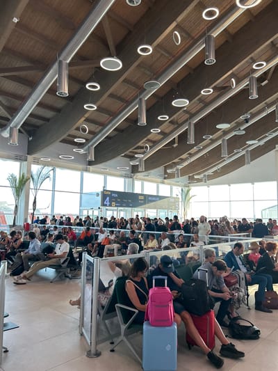 The image shows an indoor scene, likely an airport waiting area. A large group of people are seated, facing away from the camera, in a row of chairs. Many of them are looking at their phones or talking to each other. Luggage is visible around the people, including suitcases, backpacks, and duffel bags. The setting appears to be modern, with metal and wood architectural elements and circular light fixtures. Large windows line the back of the waiting area, allowing natural light to enter. In the distance, palm trees are visible through the windows, suggesting a warm climate. A departure board displays flight information, including destinations such as "BRUXELLES" and "PISA", and gate numbers like "4". The overall atmosphere seems to be one of waiting, with a sense of calm and anticipation. The lighting suggests it is daytime.