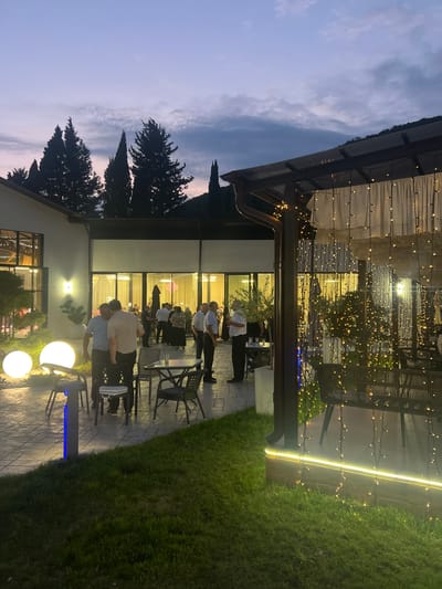 In Achadara, Abkhazia, at dusk, a group of men in casual attire and what appear to be uniformed staff stand around tables on an outdoor patio. The patio is paved with interlocking stones and features several large, spherical lights. To the right, a covered seating area is adorned with numerous string lights, creating a festive atmosphere. Beyond the patio, a building with large glass windows is visible, and trees line the background. The sky displays a soft gradient of purple and blue, with scattered clouds. The overall scene suggests an outdoor gathering or event.