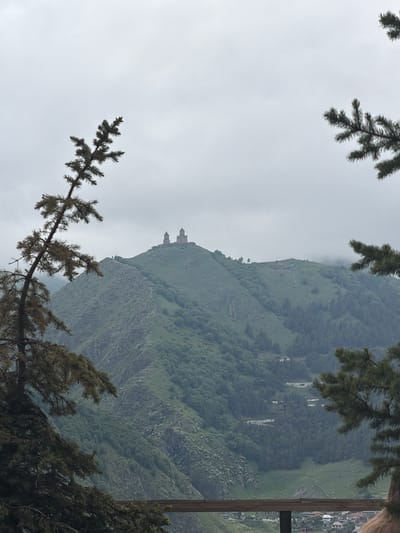 A mountain landscape with the Gergeti Trinity Church visible on a peak. The church has two domes and is made of stone. The mountain is green and covered in trees, with a road winding up its side. The sky is overcast and gray. In the foreground, there are two large evergreen trees framing the scene. Below the mountain, a small village can be seen with red-roofed houses. The overall impression is one of a remote and majestic natural setting.