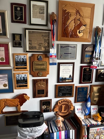 The image shows a wall covered with framed awards, plaques, and medals, predominantly associated with the Lions Club. A large wooden carving of a lion's head is a prominent feature. There are also several framed photographs and certificates. Below the wall decorations, there is a black paper shredder with a white plastic bag underneath, and stacks of binders and folders on a shelf. Small stuffed lion toys are placed among the binders. A small blue flag with a Lions Club logo is also visible on the shelf. The setting appears to be a home or office decorated with memorabilia. The text on some of the awards and documents indicates a connection to "Lions Club," "Leader Dogs for the Blind," and "Life Membership" for an individual named "Robert W. Jenkins." The location of this scene is in Sun City Center, United States of America.