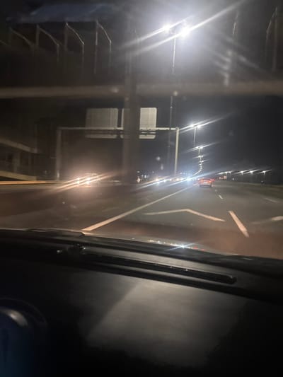 A nighttime scene shot from inside a vehicle. The view is partially obstructed by the car frame on the right and a vertical structure with horizontal bars to the left, possibly scaffolding. A brightly lit road stretches into the distance, flanked by streetlights and a row of vehicles. The road has white line markings. A building can be seen behind the scaffolding. The overall impression is a dark, urban setting seen through a reflective surface.