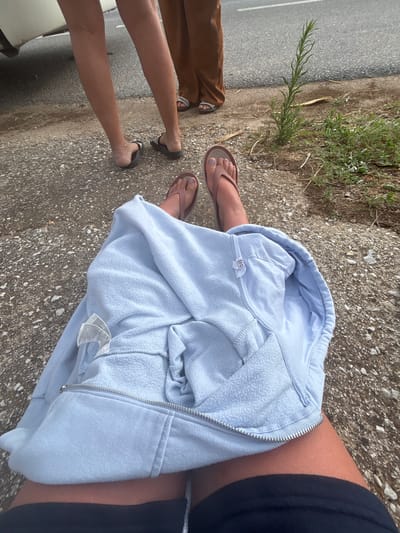 The image shows the legs of at least three individuals sitting on a gravelly surface adjacent to a paved road. One person in the foreground is wearing a light blue hooded jacket and dark shorts, with their legs crossed. Their feet are adorned with pink flip-flops. To their left, another individual is seated and partially visible, also wearing flip-flops. In the background, two other individuals are standing. One is wearing brown pants and white sandals, while the other's legs are visible in dark sandals. The environment appears to be outdoors, with a stretch of asphalt road and a patch of dry grass and weeds to the right. The location is the city of Prosotsani Municipal Unit, Greece.
