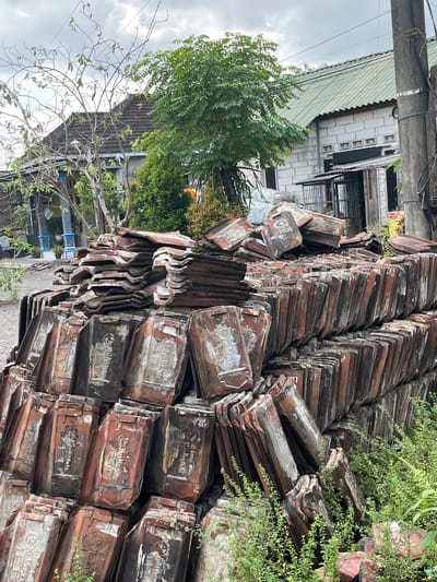 In Jati Alun Alun, East Java, a large pile of old, weathered terracotta roof tiles is stacked outdoors. The tiles are predominantly reddish-brown, with some showing signs of wear and staining. Many of the tiles in the foreground feature embossed designs, appearing to be a crest or emblem with intricate detailing. The tiles are arranged in a haphazard manner, forming a substantial mound. Behind the pile of tiles, there is a building with a green corrugated roof and white brick walls. Trees and foliage are visible in the background, and utility poles with overhead wires are present on the right side of the image. The sky is overcast, suggesting a cloudy day. No people are visible in the scene.