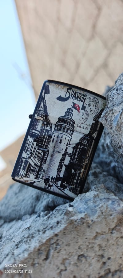 The image shows a close-up of a Zippo lighter, which is decorated with a black and white illustration of the Galata Tower in Istanbul, Turkey, and the text "HATIRASI ISTANBUL." A small Turkish flag is depicted on the tower. The lighter is resting on a rough, grey, rocky surface, possibly part of a wall or natural formation. In the background, a pale, out-of-focus sky and a textured, light-colored surface are visible. The image has a timestamp overlay in the bottom left corner that reads "GROZA_PRO, 2026/06/21 11:23." The scene is not located in the city of Истиқлолият, Tajikistan.
