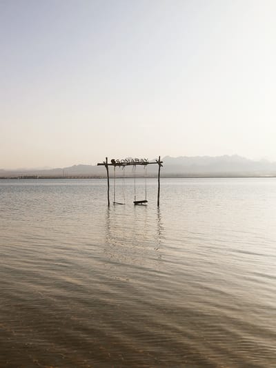 A long shot depicts a swing set with the name "SOMABAY" suspended in the ocean, with the city of Ras Abu Soma, Egypt, visible in the background. The swings are simple, made of wood, and hang from ropes tied to the crossbar of the set. The water is calm, with subtle ripples and a reflective surface. The sky is a pale, hazy color, suggesting either dawn or dusk. The coastline is visible in the distance, with buildings and low hills, giving a sense of depth to the scene. The overall atmosphere is serene and tranquil.
