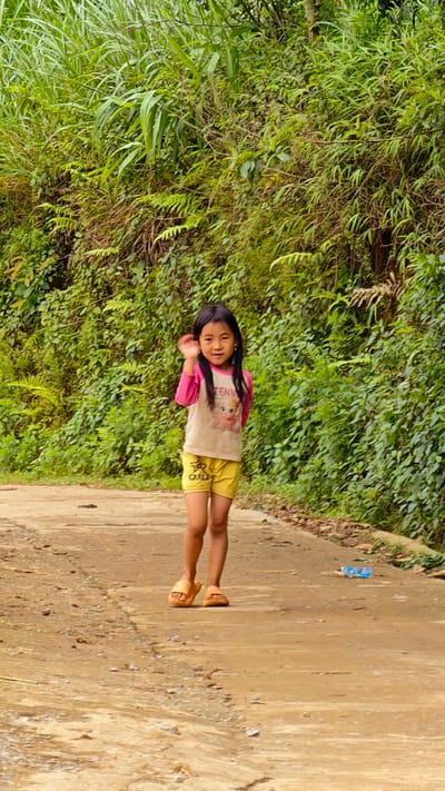A young East Asian girl, approximately 5-8 years old, stands centered on an unpaved, light brown dirt and gravel path in an outdoor setting in Pung Luong, Vietnam. She has long, straight dark hair and a light complexion, with a small earring visible in her left ear. The girl wears a long-sleeved shirt with pink sleeves and a light-colored torso featuring a partially visible graphic of a cartoon character (possibly "Elsa" from Frozen) and text "TENW". She is also wearing yellow shorts with dark markings on the left leg and orange open-toed slide sandals. Her right arm is raised, with her hand open, indicating a wave towards the viewer. She maintains direct eye contact with a slight smile. The background consists of a dense, vibrant green wall of diverse foliage, including tall grasses, ferns, and various leafy plants, suggesting a lush, natural environment. On the far right edge of the path, a small blue plastic bottle is visible. The path itself shows scattered small stones and an uneven texture.