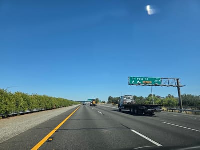 A flatbed truck drives on the right lane of a multi-lane highway under a bright, clear blue sky. The truck, with a white cab and a dark flatbed, is the most prominent object. In the distance, a yellow excavator is visible on the left side of the highway. A green overhead highway sign indicates "Del Paso Rd EXIT 525A" with an arrow pointing left. The highway is lined with trees and bushes on the left and right sides. The road surface is asphalt with clear lane markings. The scene appears to be on a sunny day, likely in the city of Sacramento, California, United States of America.