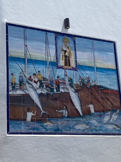 This is a detailed description of a ceramic mural located in Conil de la Frontera, Spain, depicting a scene related to fishing and religious veneration.
The mural is composed of numerous ceramic tiles, arranged in a grid pattern and framed by a dark blue border. The artwork portrays a bustling fishing scene at sea, with a central focus on several large fish, possibly tuna, being brought aboard a boat. Several men, dressed in simple, historical-looking clothing, are actively engaged in the fishing operation. They are shown pulling ropes, handling the fish, and standing on the deck of the boat. The water is depicted with varying shades of blue, suggesting depth and movement.
Above the fishing scene, within a smaller, ornate frame, is an image of a religious figure, likely the Virgin Mary, holding a child. This religious iconography is placed centrally in the upper portion of the mural, implying a connection between the fishing activities and a patron saint or religious blessing. The background of the larger mural features a horizon line where the sea meets a pale sky with subtle color gradations.
In the foreground, below the main deck of the boat, some figures are partially submerged in the water, possibly fishermen or figures associated with the sea. More fish are visible swimming in the water. The overall aesthetic of the mural suggests a narrative of the town's maritime heritage and its connection to religious faith.
The mural is mounted on a textured white wall. A dark, cylindrical light fixture is mounted on the wall above and slightly to the left of the mural. The texture of the wall is rough and irregular, providing a contrast to the smooth surface of the tiled artwork.