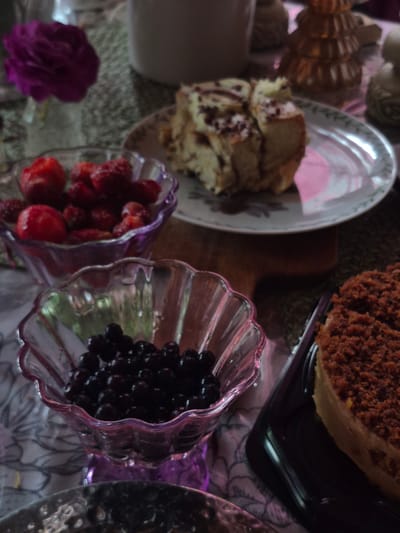 The image depicts a close-up, overhead view of a table setting featuring desserts and fruit. In the foreground, a clear glass bowl filled with dark blueberries is visible, set against a floral patterned tablecloth. To the left, another clear glass bowl holds bright red strawberries. Behind these bowls, a slice of cake with chocolate frosting sits on a patterned plate. To the right, a slice of cake with a crumb topping is presented on a black rectangular platter. A single purple rose in a vase is situated in the upper left corner of the frame. Decorative elements, including what appears to be a golden Christmas tree-shaped candle holder, are also present in the background. The lighting is dim, creating a moody atmosphere. The scene is set indoors. The specific location in Астра-1 Днт, Russia is not discernible from the image itself.