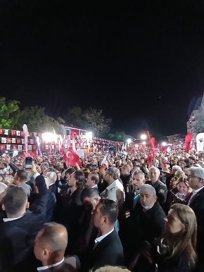 The image shows a large crowd of people gathered in Silivri, Türkiye, at night. The crowd is densely packed, with many people holding red flags with a white crescent moon and star. Some people are holding cell phones up, possibly taking pictures or videos. The sky is dark, and streetlights illuminate the crowd. Some people are looking at something in the distance, while others are talking to each other. There are trees and buildings in the background. The location of the image is determined by the coordinates 41.071 latitude and 28.244 longitude. The scene appears to be a political rally or a public gathering.