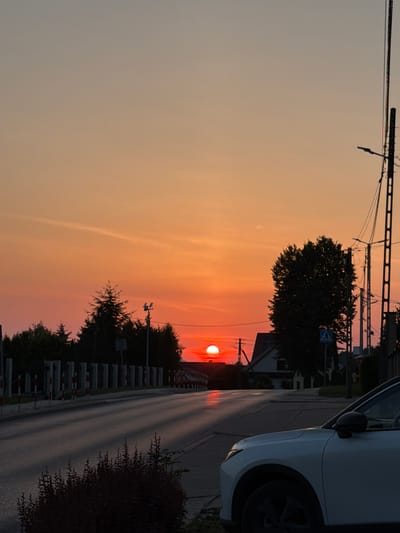 The image depicts a street scene at sunset in Mordarka, Poland. The sky is a gradient of orange and pink, with the sun low on the horizon, casting a warm glow. A white car is parked on the right side of the road, partially in the foreground. The road itself stretches into the distance, lined with trees and streetlights. A building with a dark roof is visible behind a cluster of trees on the right. The left side of the road features a barrier with red and white markings and a row of trees. The overall impression is a peaceful, rural evening setting.
