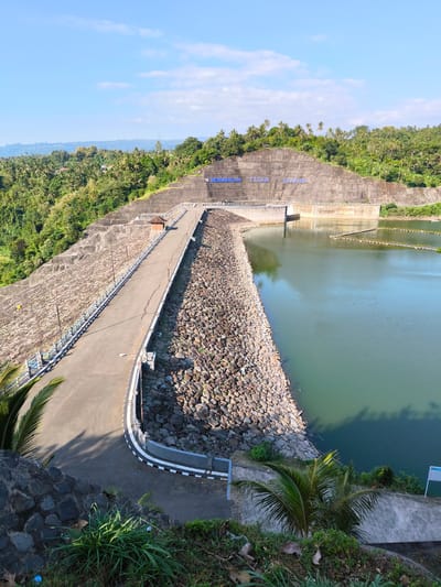 This image captures the Titab-Ularan Dam in Seririt, Indonesia, under a clear blue sky with sparse white clouds. The central feature is a large, grey concrete dam wall with the name "BENDUNGAN TITAB - ULARAN" prominently displayed in blue capital letters across its upper face. To the right of the dam, a calm, greenish-blue body of water, likely a reservoir, extends, featuring several linear floating barriers or structures. A paved asphalt road, bordered by white and blue guardrails and equipped with streetlights, curves across the top of the dam structure, separating the reservoir from a rocky, vegetated slope. A small, brown-roofed building is situated alongside this road on the left. The surrounding hills and embankments are densely covered with various green foliage, including numerous palm trees, particularly on the left side and in the distant background. The sun casts clear shadows, indicating daytime. No individuals or human activity are visible in the scene.