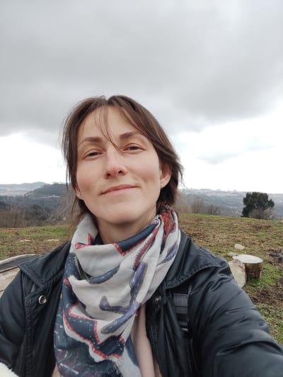 The image depicts a female subject in Bestuzhevskoye, Russia, captured in a medium close-up selfie format. The woman, appearing in her 20s to 30s, has medium-brown hair, a faint smile, and is looking directly at the camera. She wears a dark grey puffer jacket, a multi-colored scarf with blue, red, and white patterns, and a light-colored garment underneath. A small silver hoop earring is visible in her left ear. The background features an overcast sky with grey clouds, occupying the upper third of the frame. The subject is positioned on an elevated, uneven terrain covered with sparse, dry grass and patches of bare earth. Several cut wooden stumps are visible on the ground to the subject's right. In the mid-ground, a valley opens to a densely populated urban area or city, characterized by numerous buildings and structures, against a backdrop of distant hills and mountains. Scattered leafless trees and a prominent dark green conifer tree are also visible in the mid-ground.